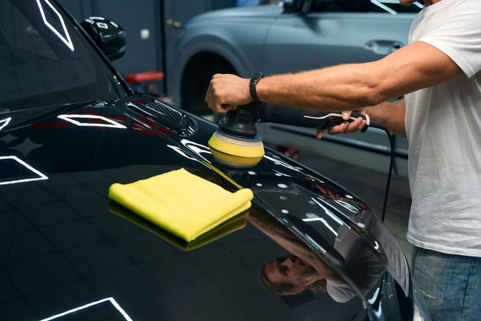 Person polishing a black car with a buffer and yellow cloth in a garage