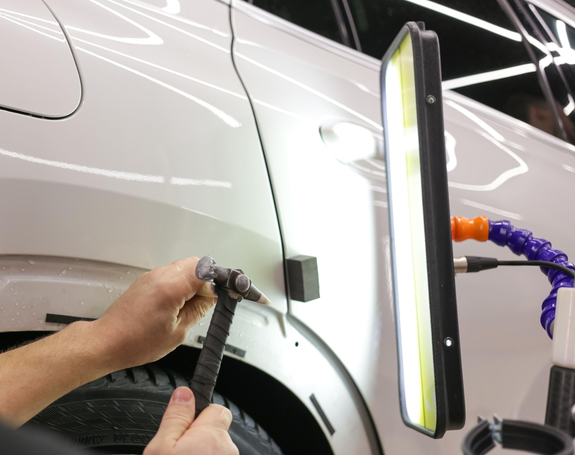 Technician repairs a white car’s rear wheel arch with a heated dent tool.