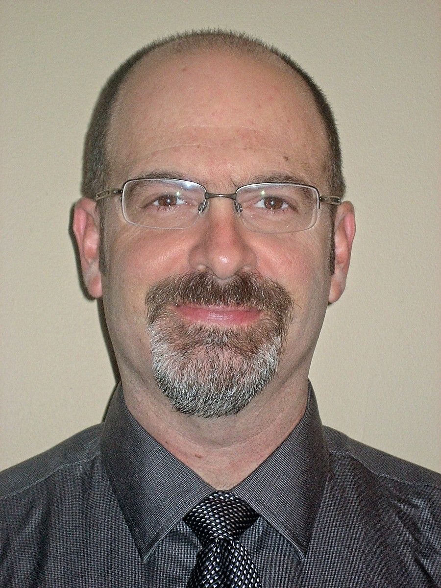 Middle-aged man with glasses, goatee, and collared shirt, smiles slightly at the camera.
