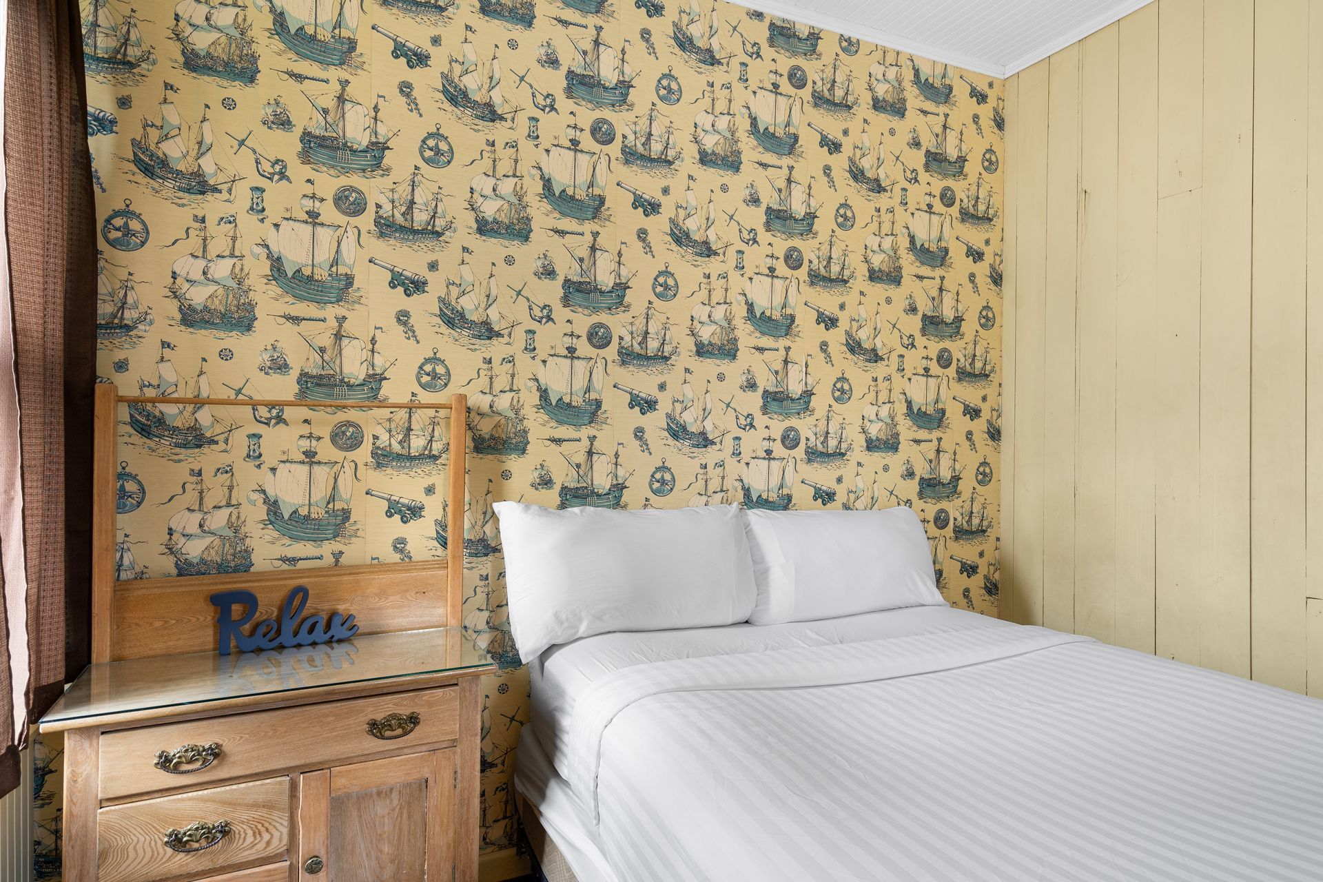A bedroom with a bed , nightstand , and wallpaper with ships on it.