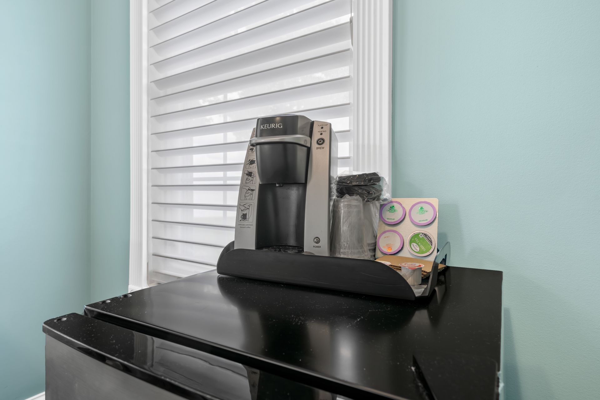 A coffee maker is sitting on top of a black refrigerator next to a window.