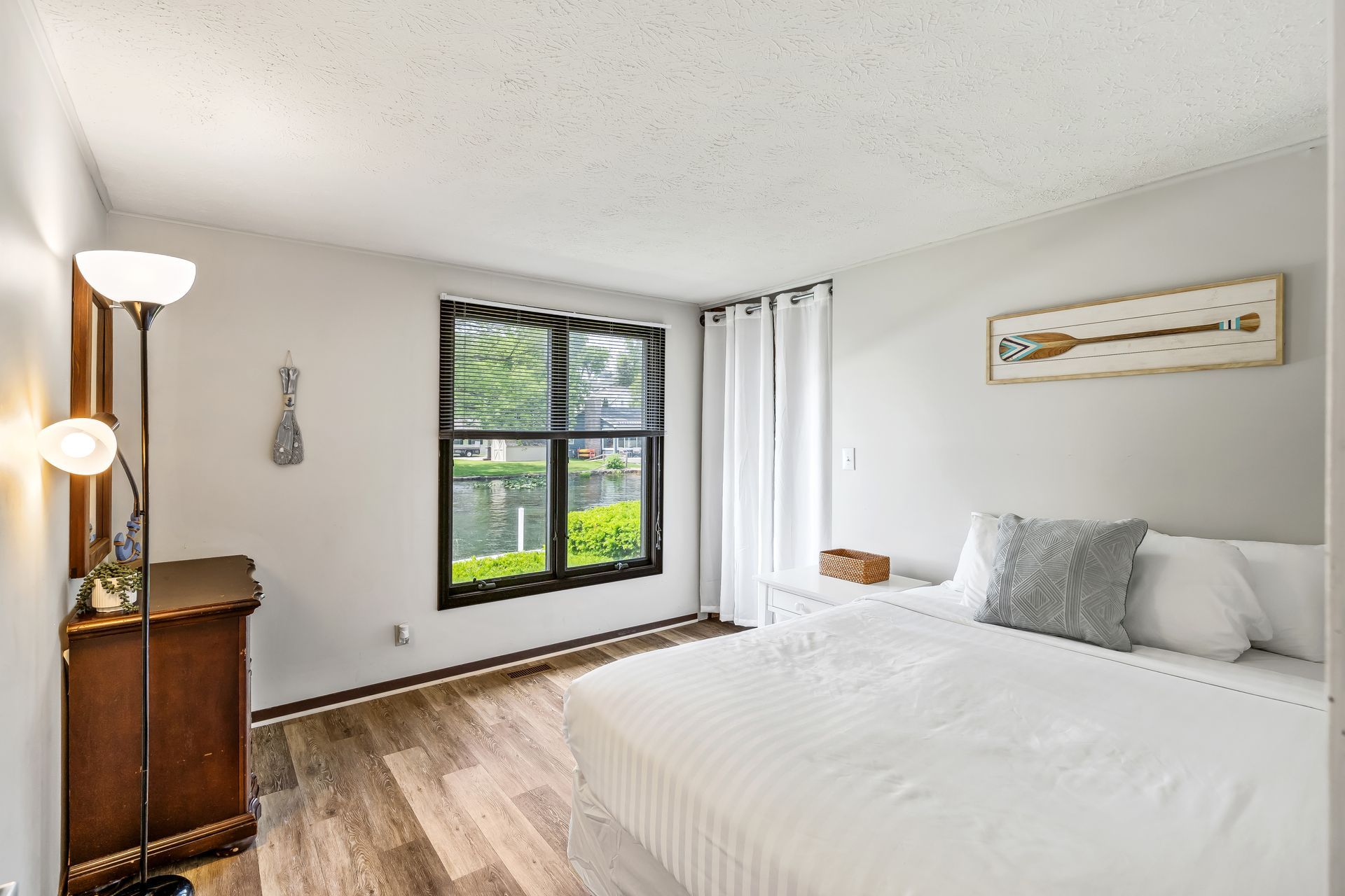 A bedroom with a bed , dresser , lamp and two windows.