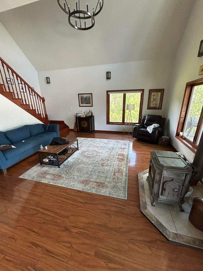Bright living room with blue sofa, patterned rug, staircase, fireplace, and large windows on hardwood floors