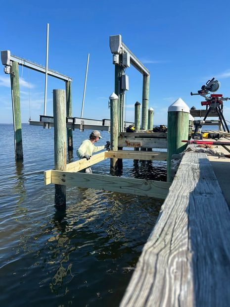 Person working on a wooden dock pile in bright blue waterfront setting, with boat lift and camera setup nearby