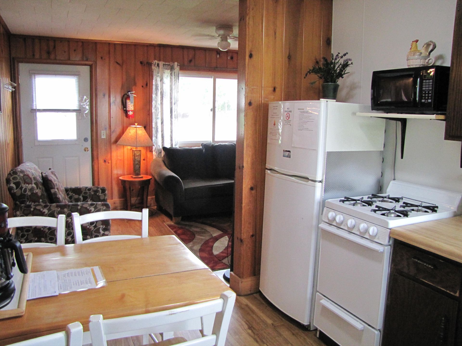 Cozy cabin interior: kitchen with stove, fridge, table, leading to living room with couch, chairs, and window.