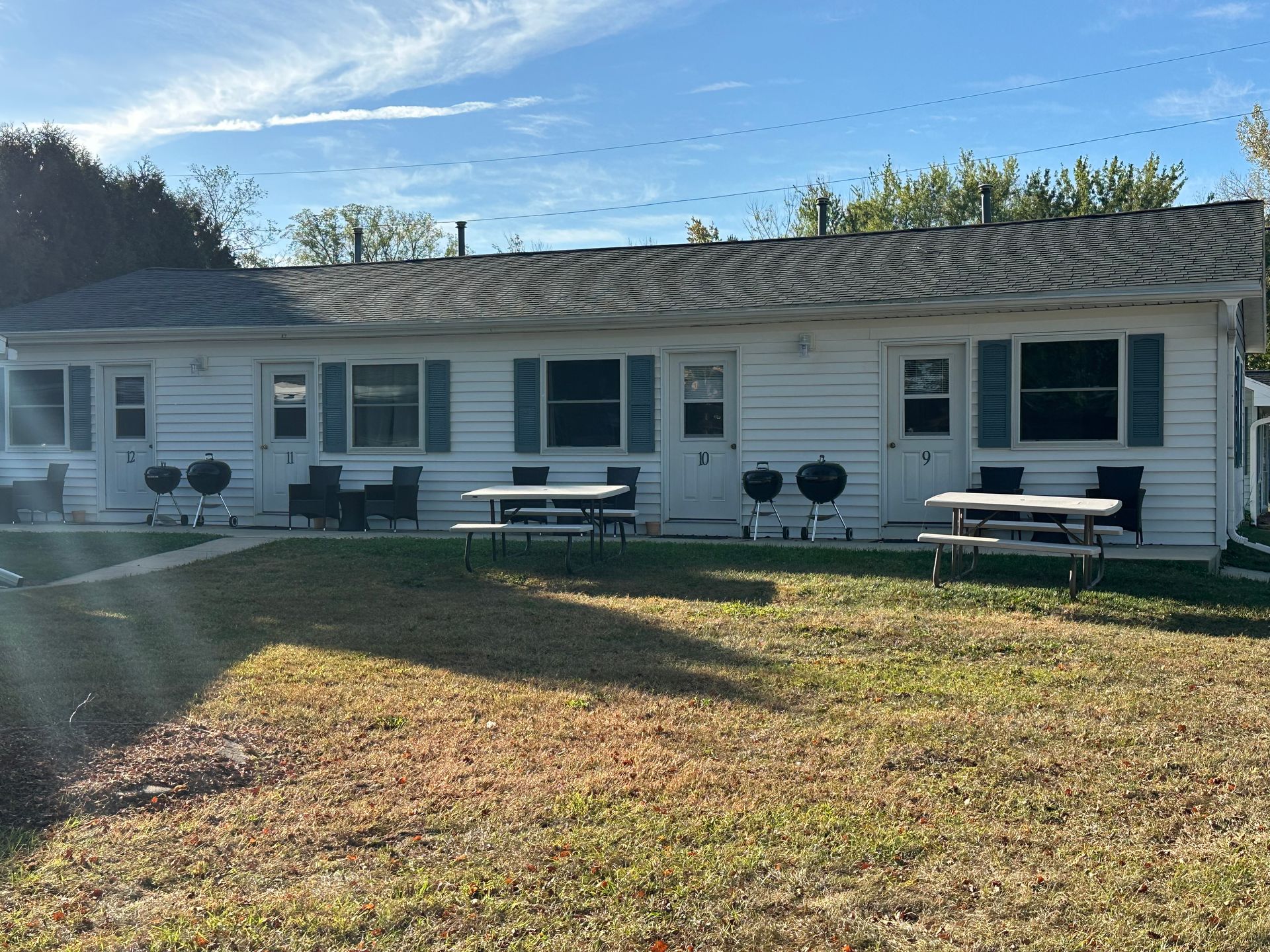 White one bedroom cabins with blue shutters, picnic tables, and grills on a grassy lawn under a blue sky.