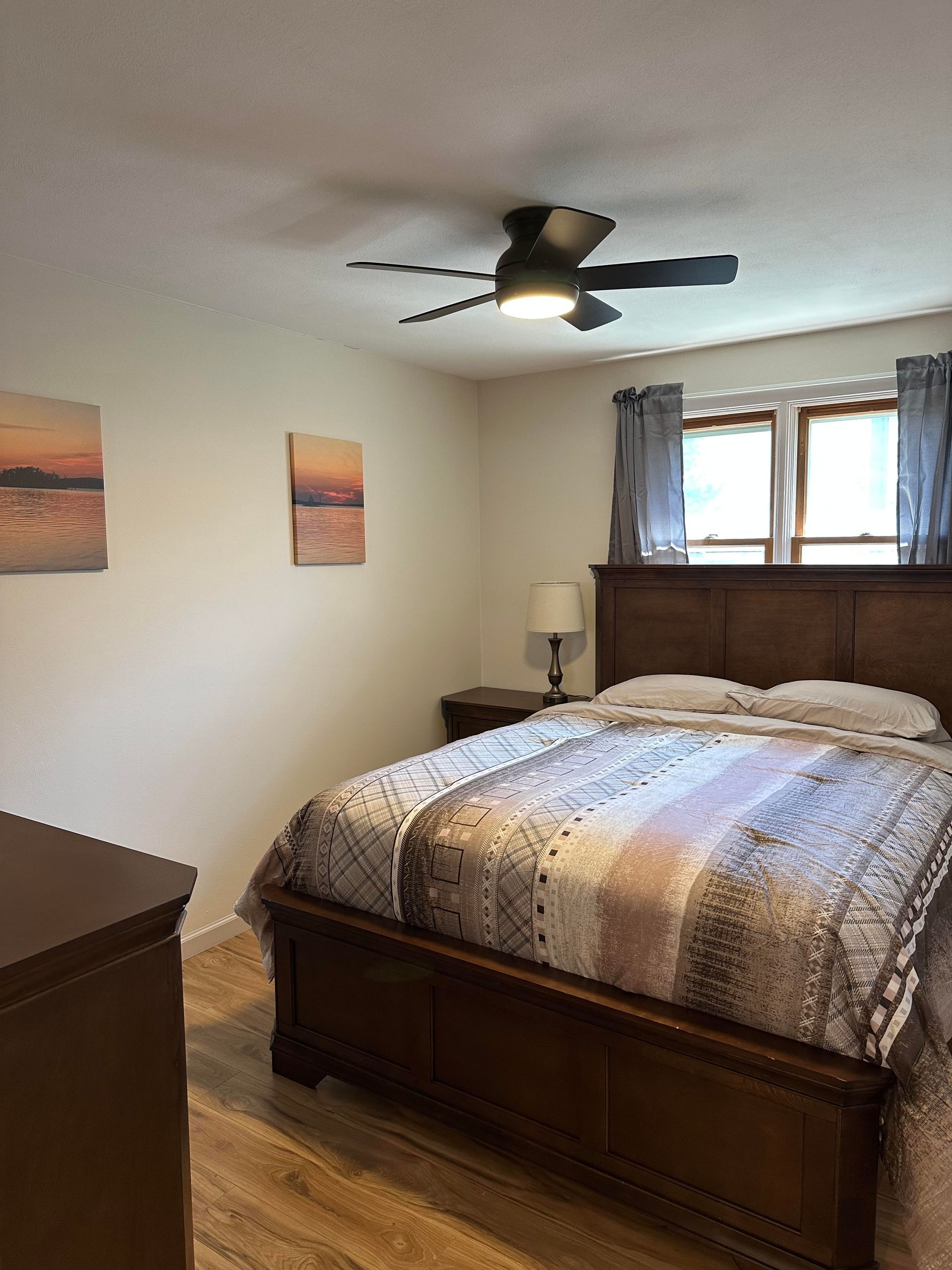 A bedroom with a wooden bed frame, a patterned quilt, a ceiling fan, and two sunset wall prints near a window.