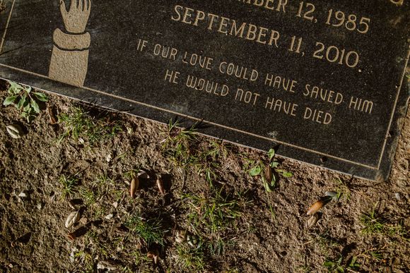 Gravestone plaque in dirt and grass, engraved with dates and the message “If God love could have saved him…”