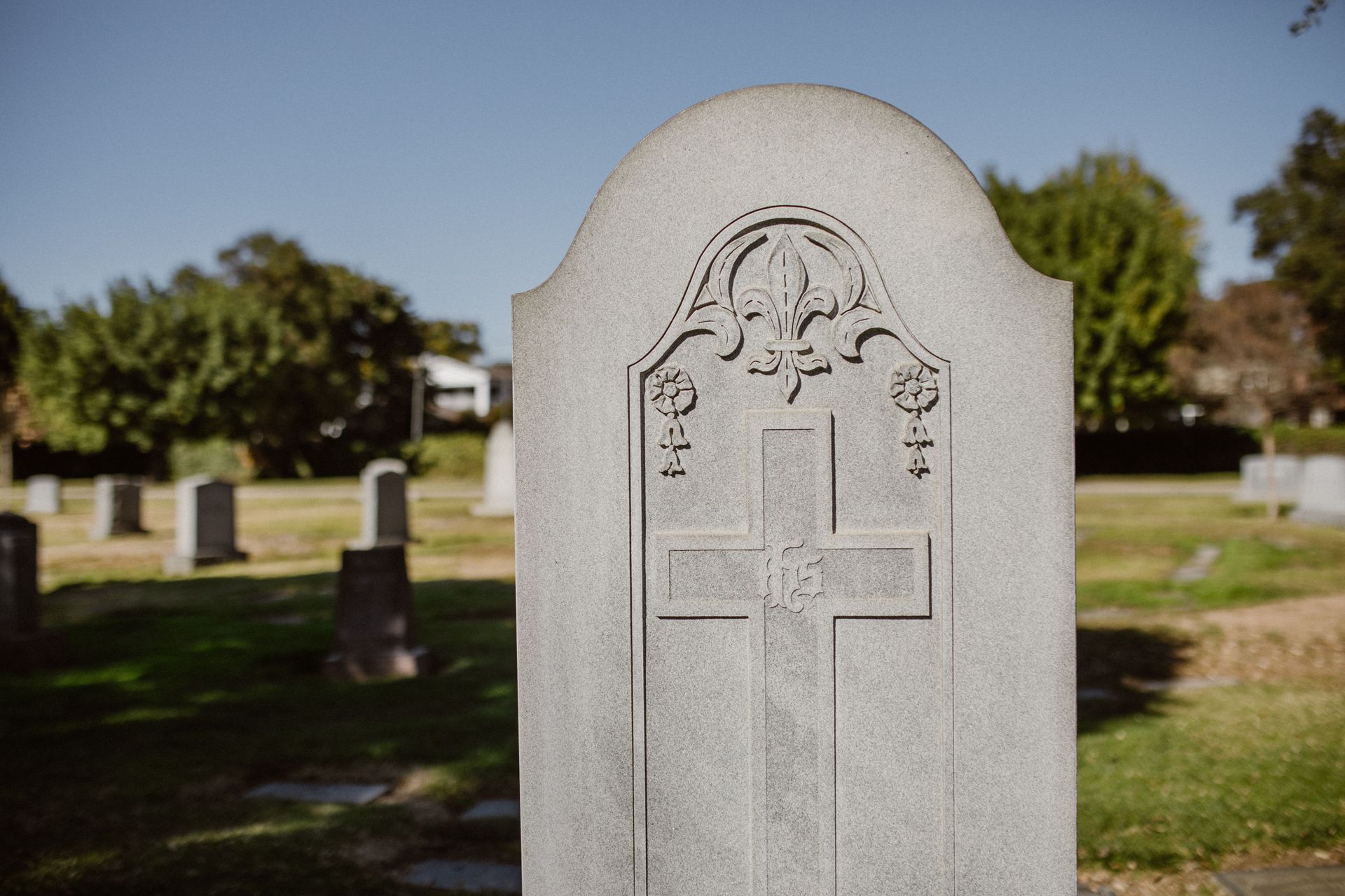 White gravestone with a carved cross in a sunny cemetery, trees and other headstones in the background