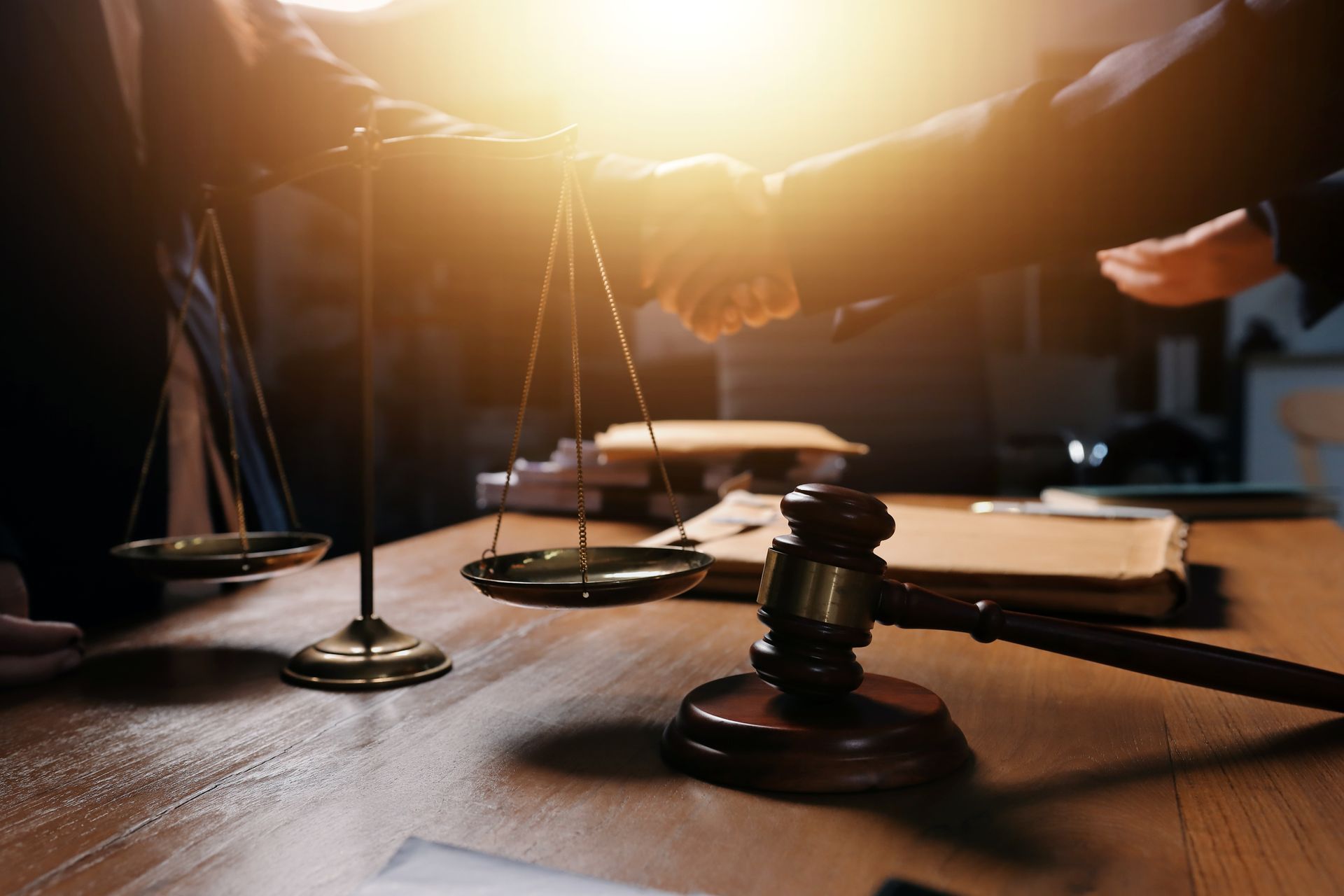 Gavel, scales of justice, and handshake on a table in a courtroom, symbolizing legal agreement.