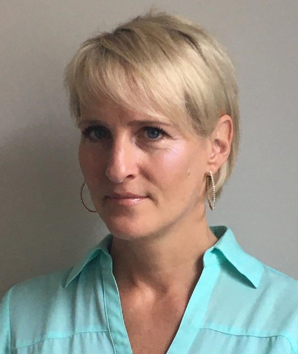 Portrait of a person with short blonde hair wearing a turquoise collared shirt, looking at the camera.