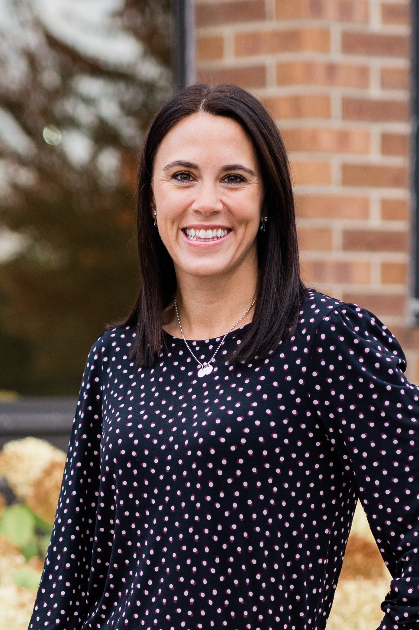 Woman in a black polka-dot blouse smiling outdoors beside a brick wall and flowers