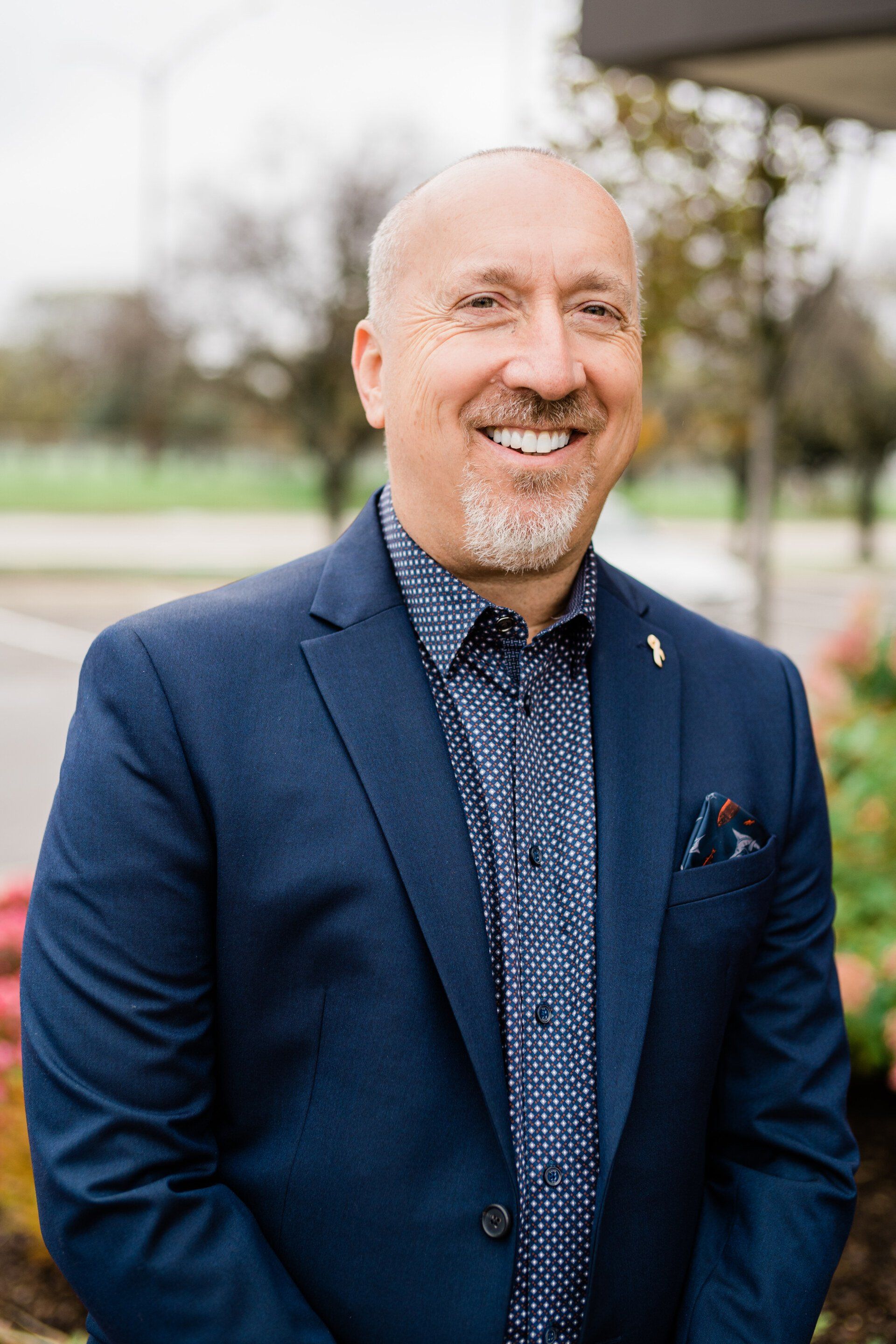 Smiling man in a navy blazer standing outdoors with a blurred park background