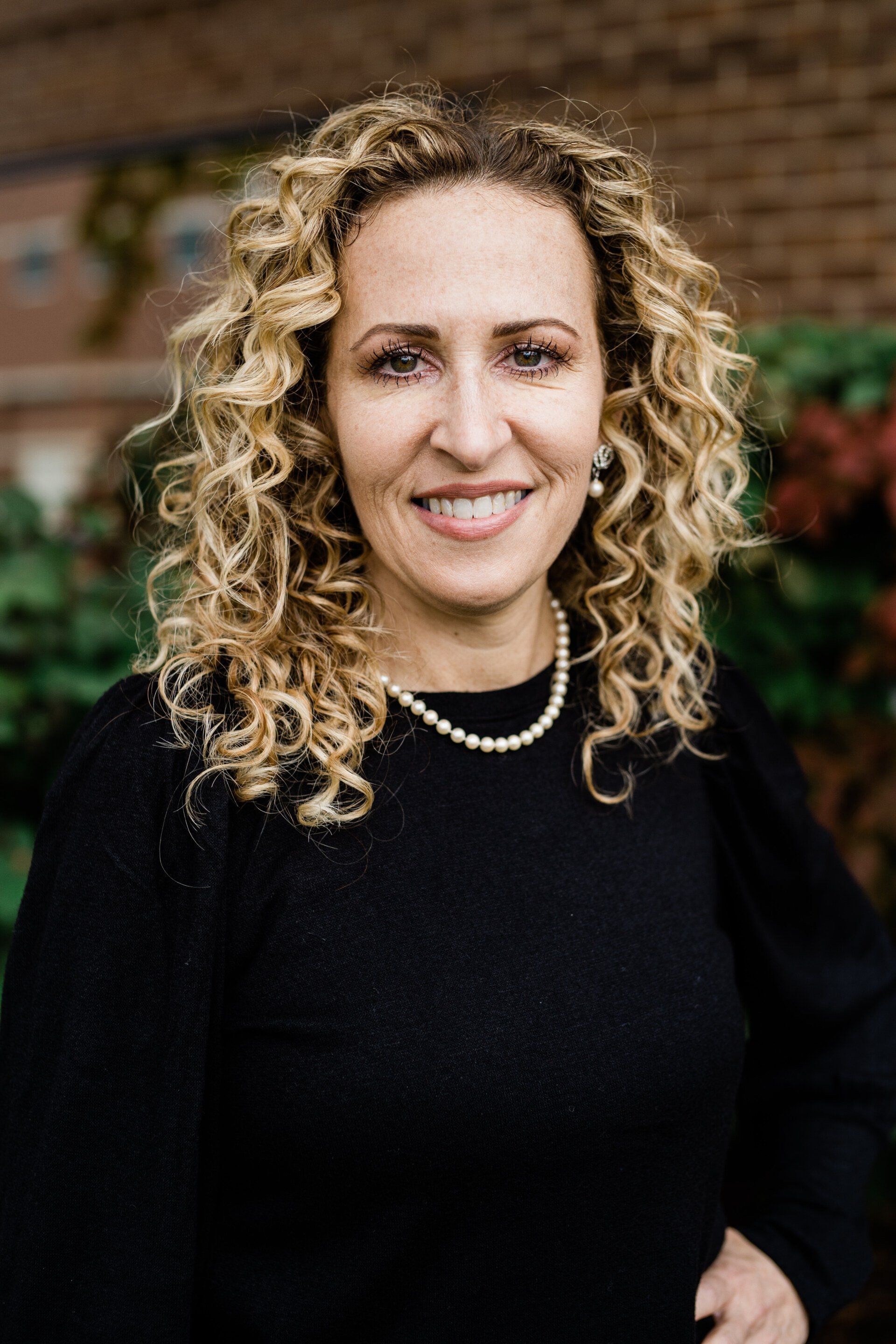 Portrait of a smiling woman with curly blonde hair wearing a black sweater and pearl necklace outdoors