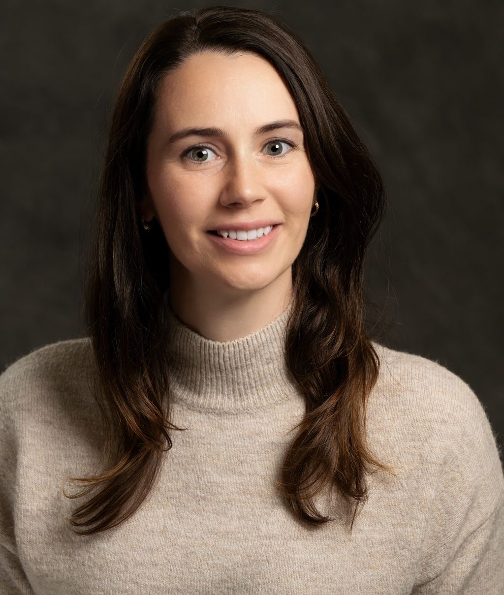 Smiling woman with long brown hair wearing a beige sweater against a dark background