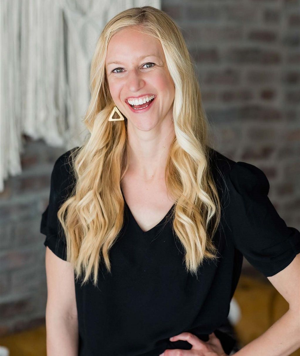 Smiling blonde woman in a black top standing indoors against a brick wall background