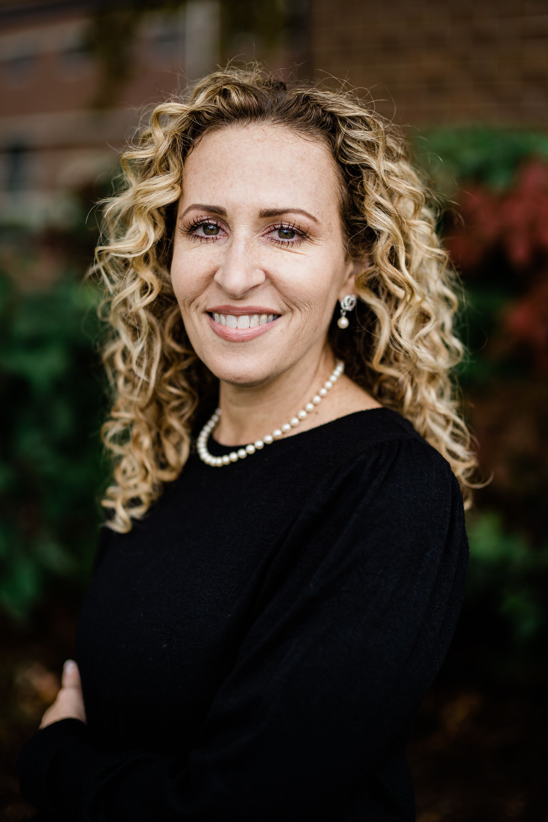 Smiling person with curly hair in a black top and pearl necklace, standing indoors against a blurred background