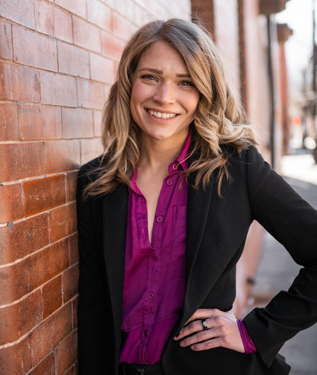 Smiling person in a black blazer and magenta blouse leaning against a brick wall outdoors