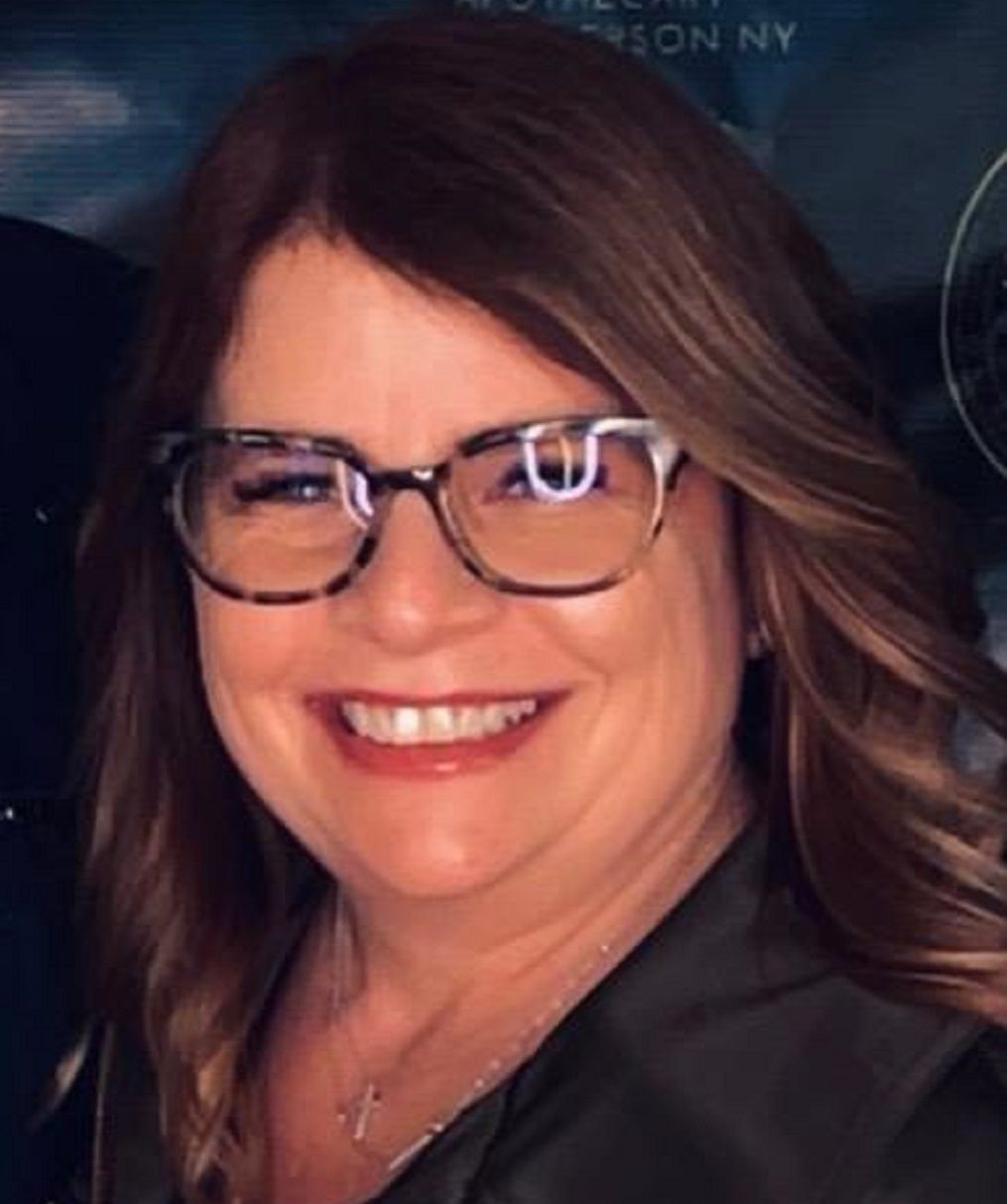Smiling woman with glasses and wavy brown hair in a dark blazer against a blue background