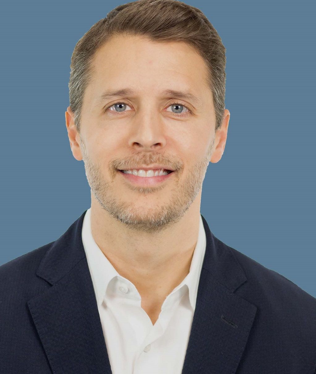 Studio headshot of a smiling man in a navy blazer and white shirt against a blue background