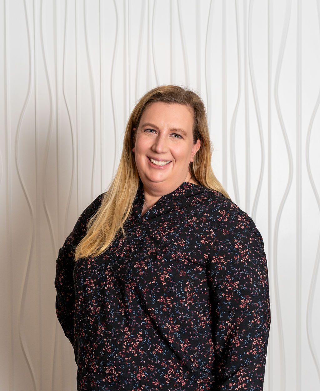 Smiling person with long blonde hair wearing a dark floral blouse against a white wavy backdrop