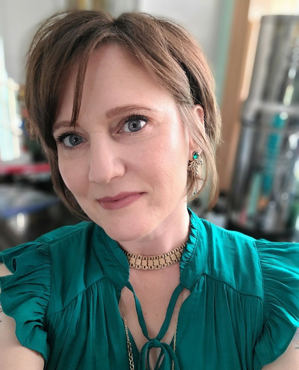 Portrait of a woman in a teal blouse, wearing a gold necklace, with a soft smile indoors.