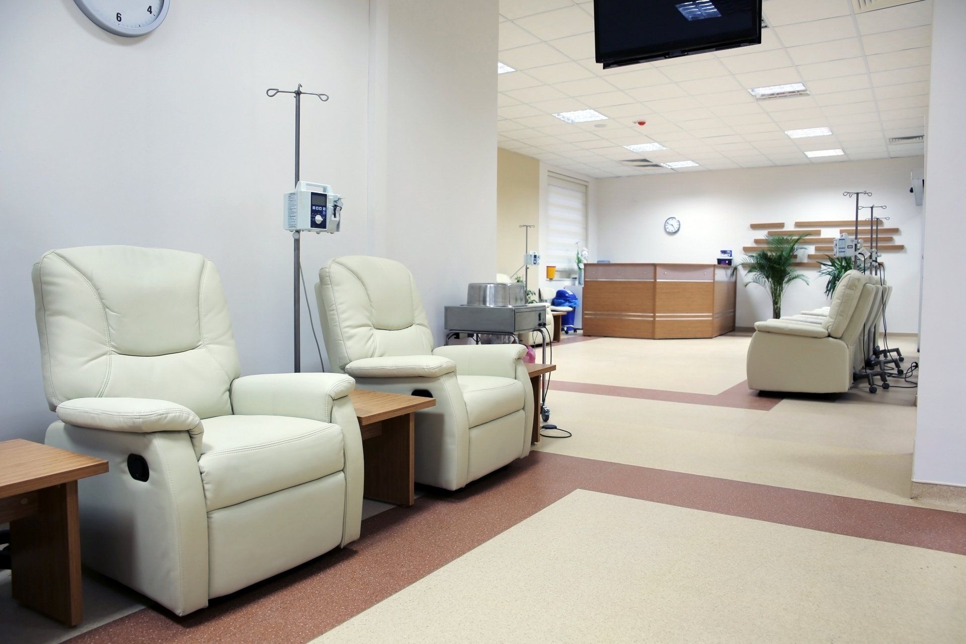 Empty medical waiting room with beige recliners, side tables, and a reception desk in the background