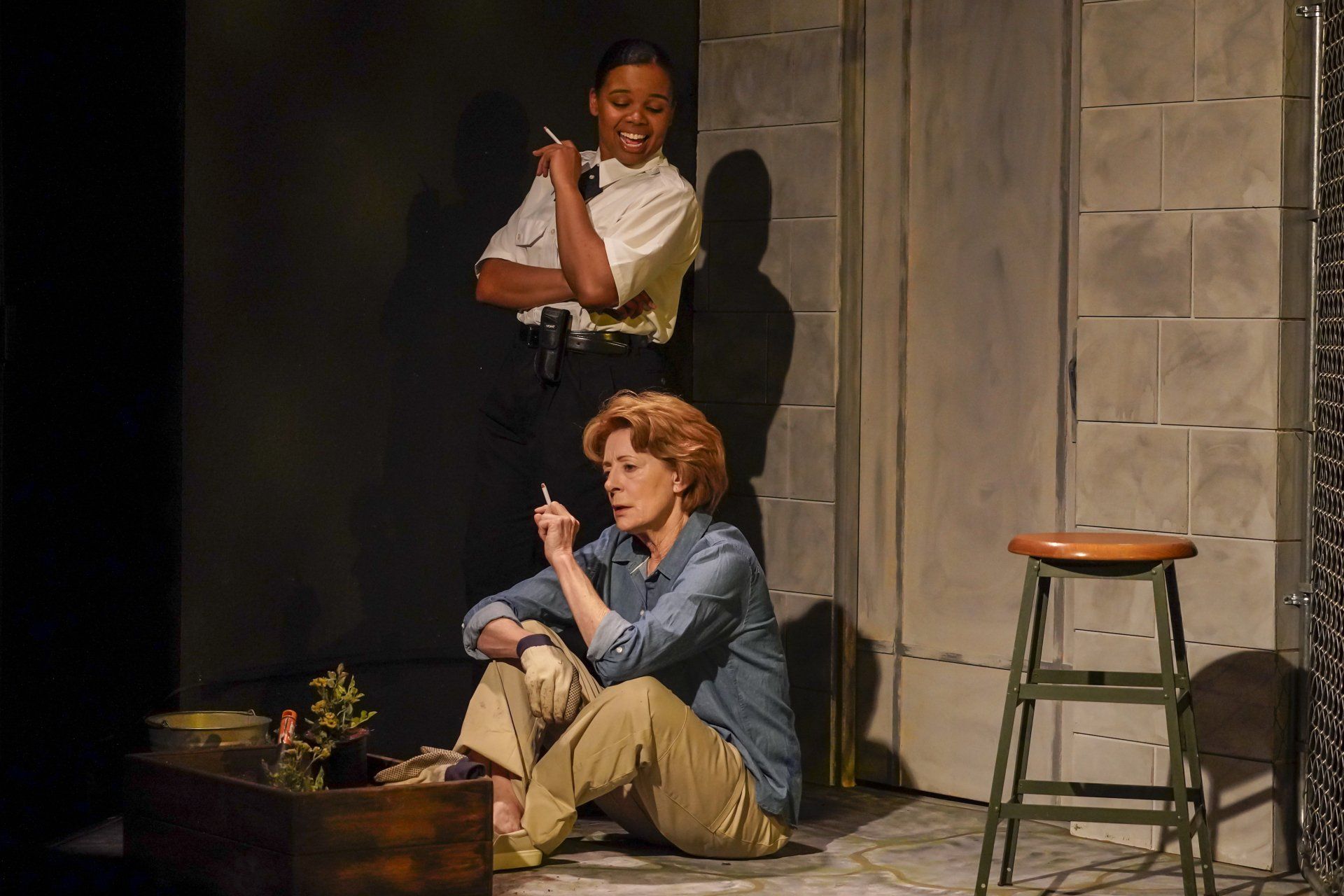 A woman is sitting on the floor while a man stands behind her while both smoke a cigarette.