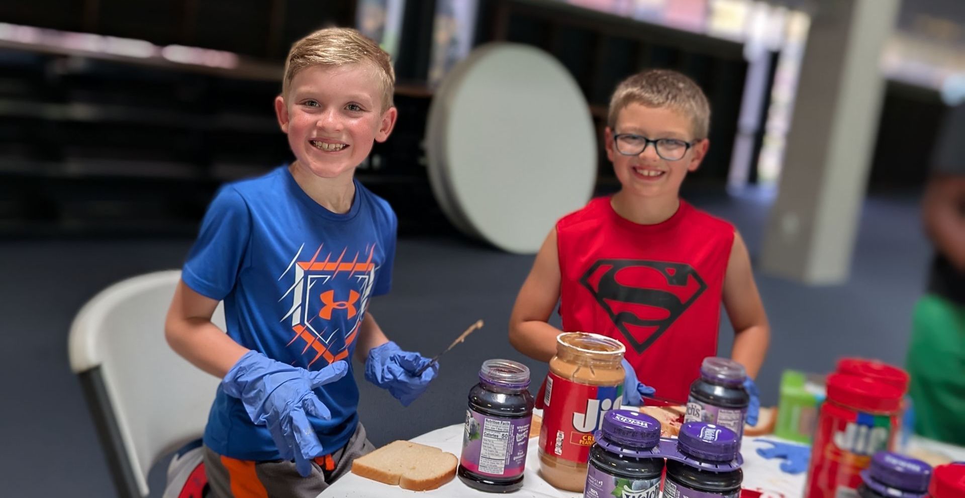Two boys making sandwiches: one in blue, one in red, smiling. Table with peanut butter, jelly, bread, and gloves.