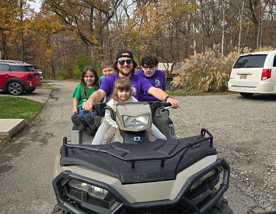 Four people ride a black ATV on a driveway, with autumn trees and parked cars in the background.