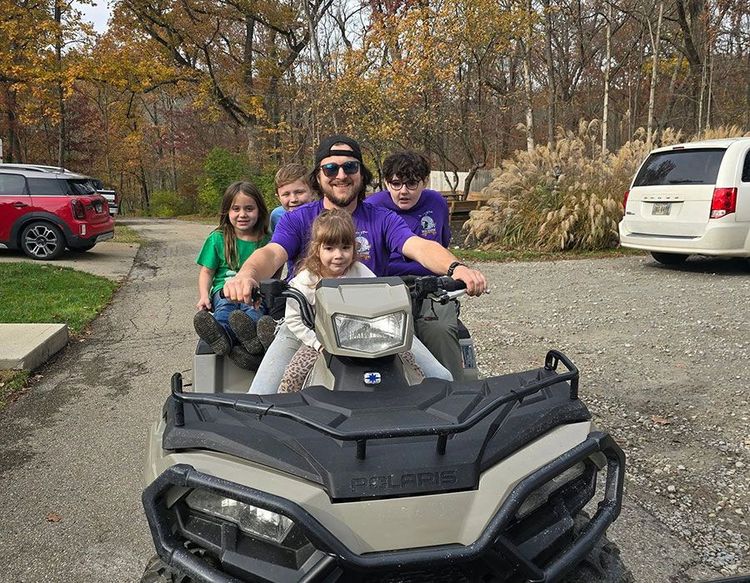 Four people ride a black ATV on a driveway, with autumn trees and parked cars in the background.