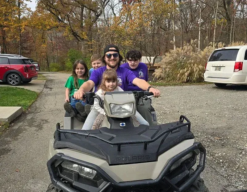 Four people riding an ATV on a tree-lined driveway, with parked cars in the background.