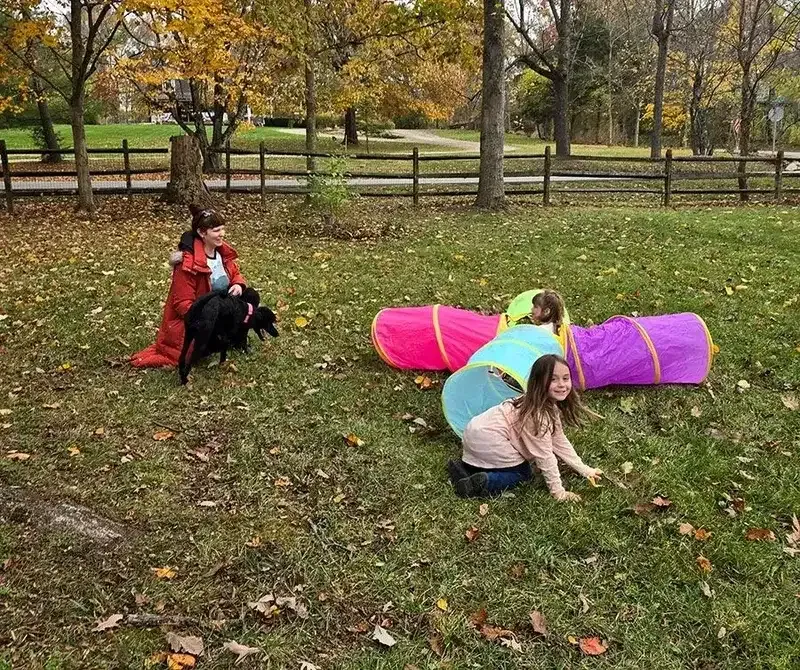 Children in colorful costumes crouch on a leaf-covered field in a park.