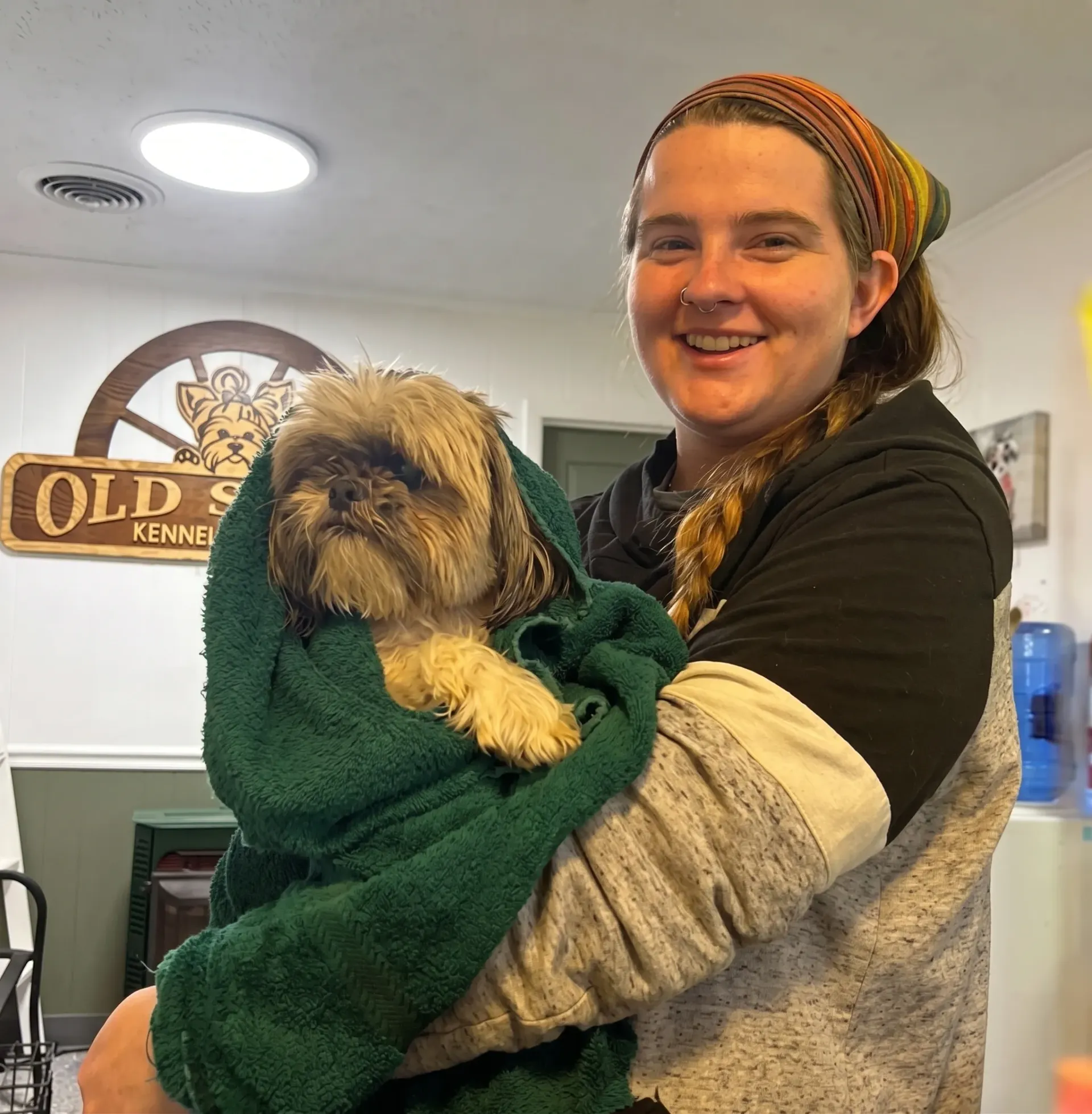 Woman holding a small dog wrapped in a green towel indoors, both facing the camera and smiling.