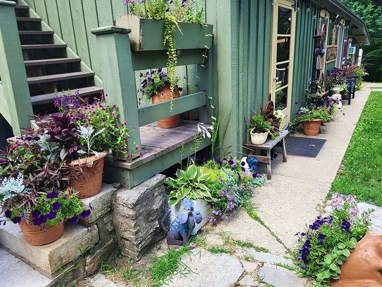 Charming garden walkway with potted flowers, trailing vines, and a green house exterior with stairs and porch.
