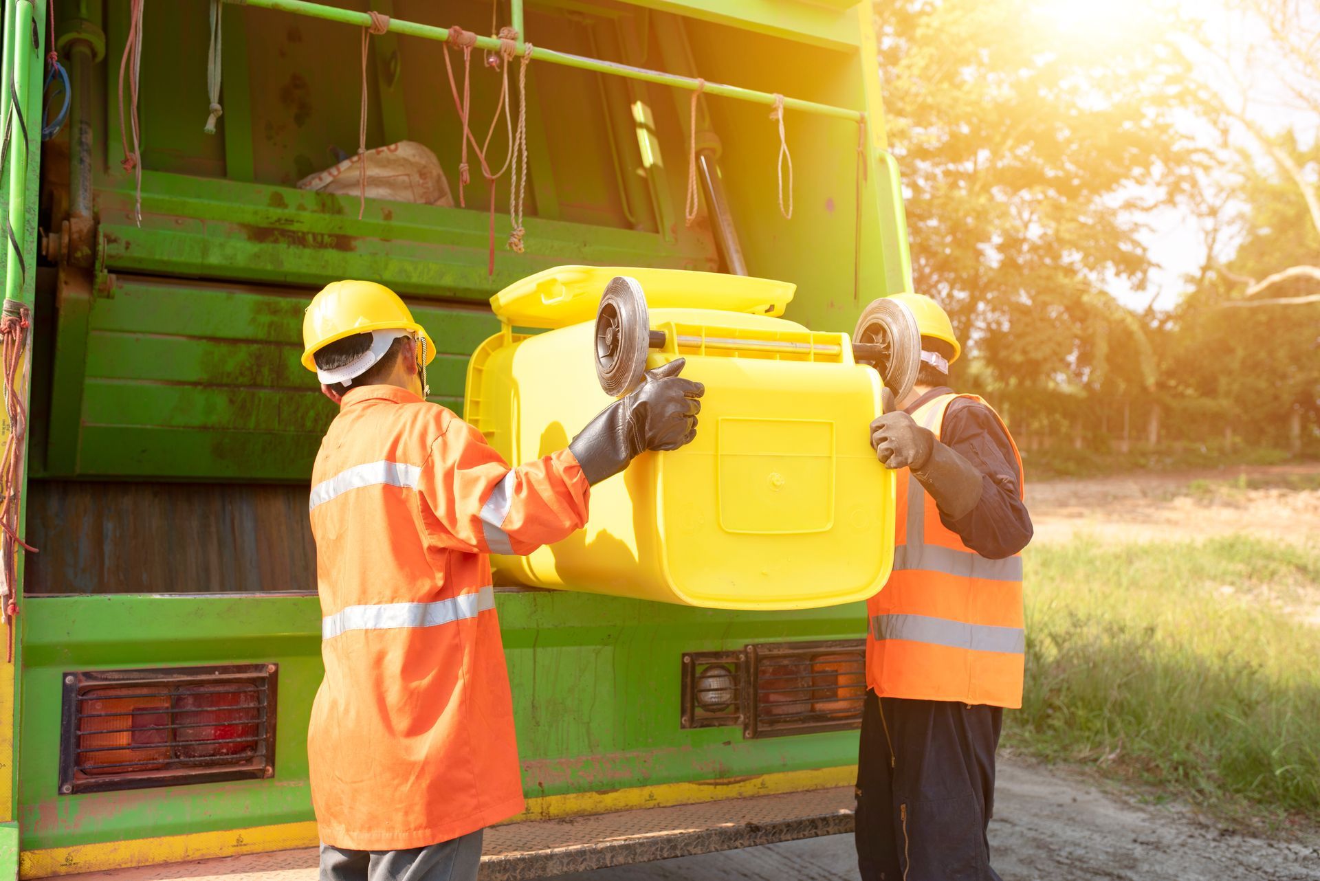 Sanitation workers wearing orange reflective vests and yellow helmets lifting a yellow wheeled trash bin into a green garbage truck. Sanitation workers wearing orange reflective vests and yellow helmets lifting a yellow wheeled trash bin into a green garbage truck.