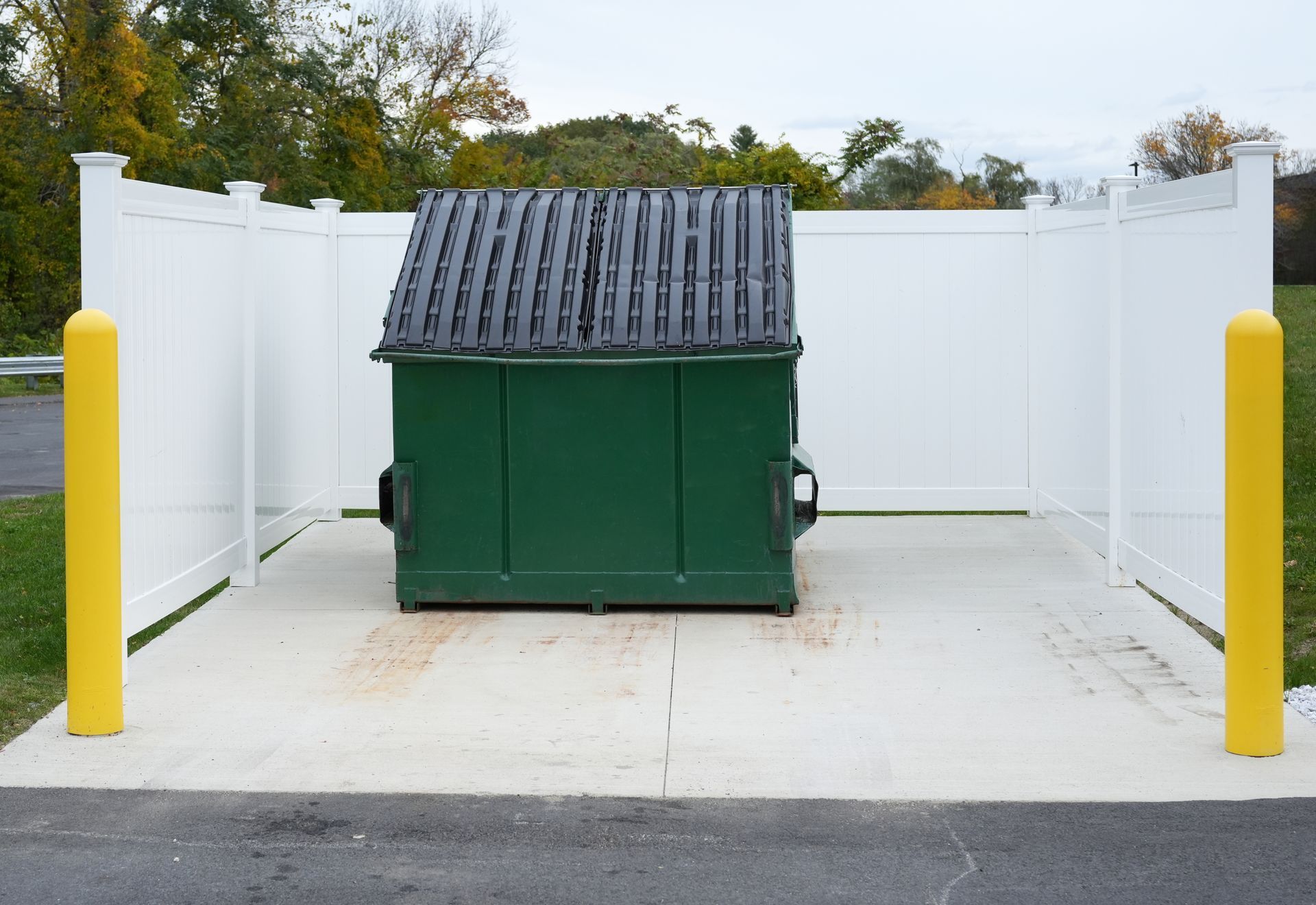 Green dumpster placed on a concrete pad, enclosed by white vinyl fencing with yellow bollards. Green dumpster placed on a concrete pad, enclosed by white vinyl fencing with yellow bollards.