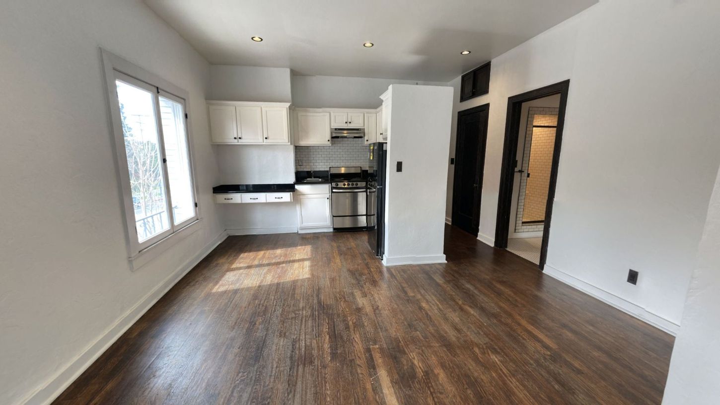 Small apartment interior with white walls, dark wood floors, kitchen, and doorway to a hallway.
