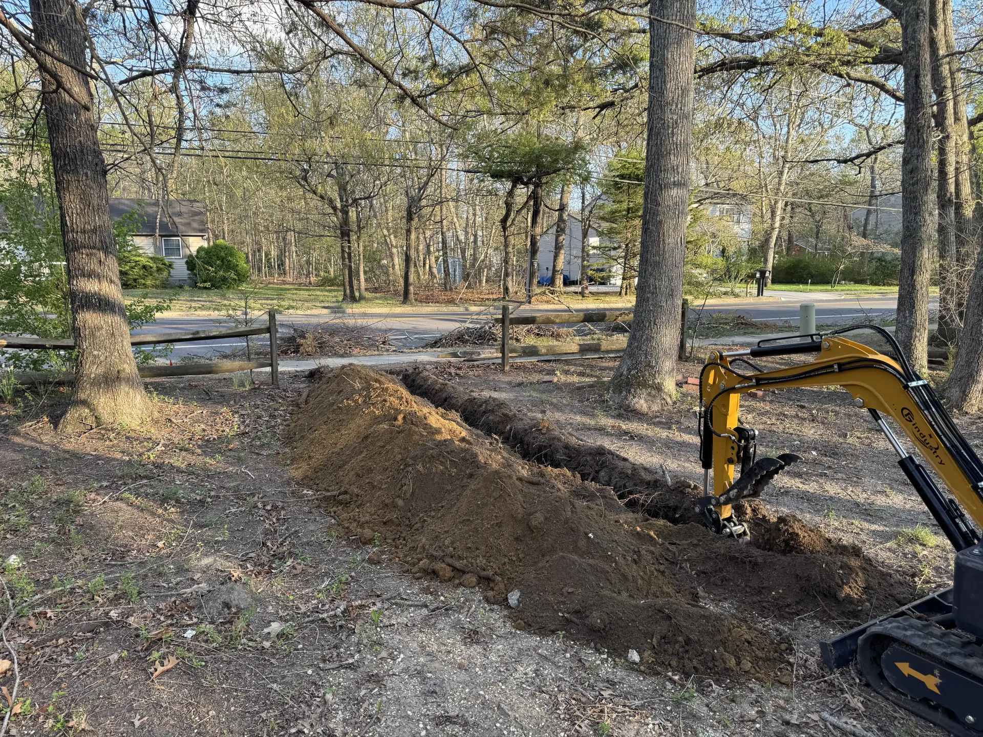 Yellow excavator digging a trench in a wooded yard, with a mound of dirt and fallen leaves around it