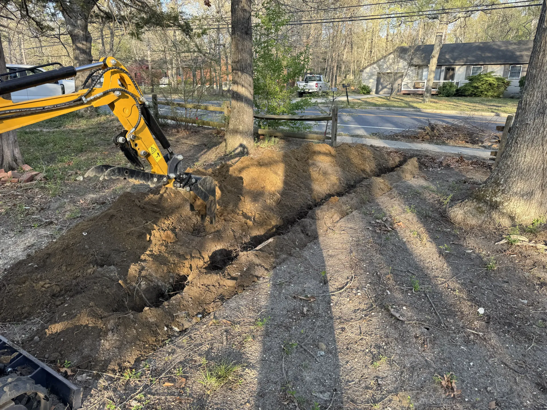 Yellow excavator digging a trench along a dirt path beside a road in a wooded area.