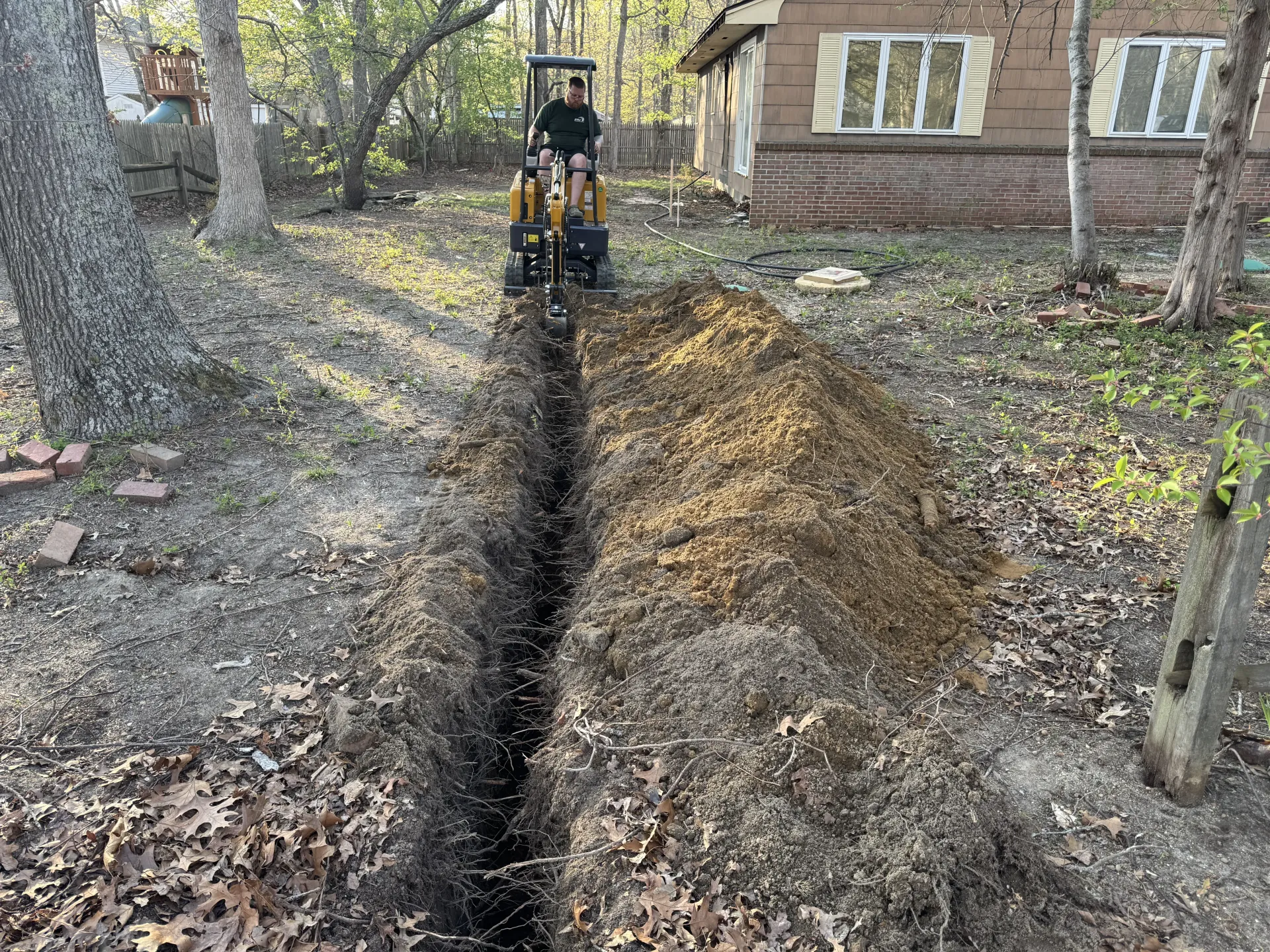 Mini excavator digging a long trench in a leafy yard beside a house