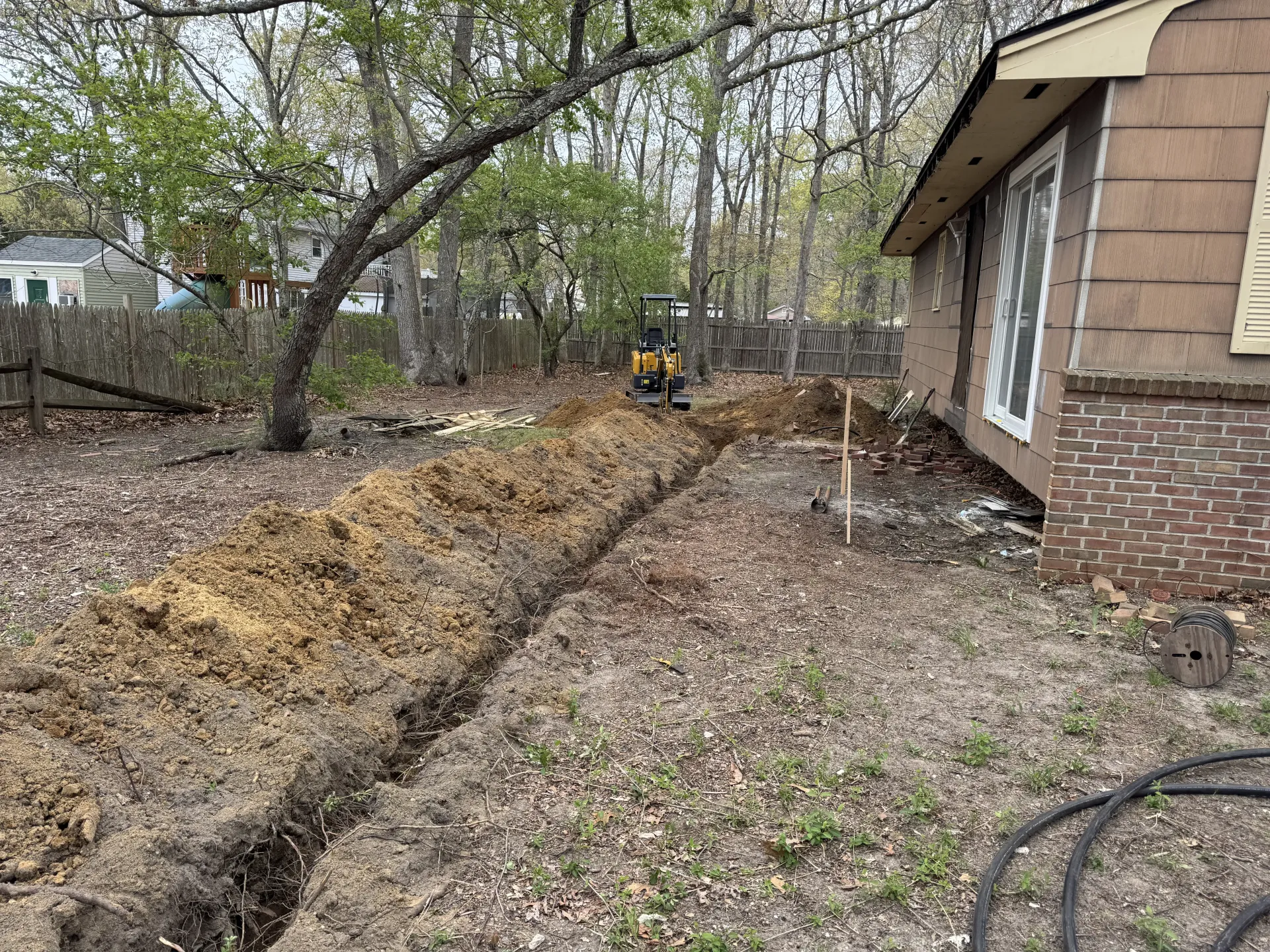Long trench dug beside a brick house in a leaf-covered yard, with trees and a backhoe in the distance