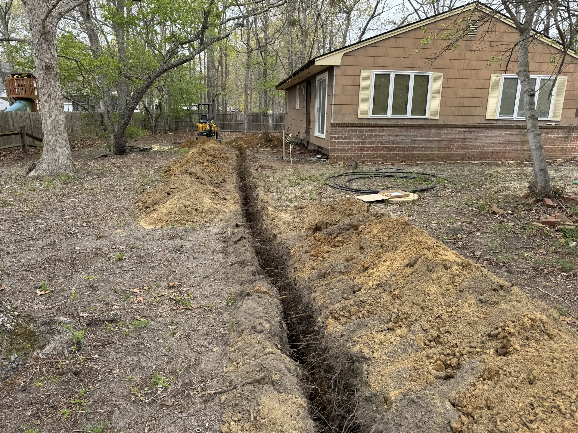 A deep trench cuts through a yard beside a brown house with trees and fallen leaves.