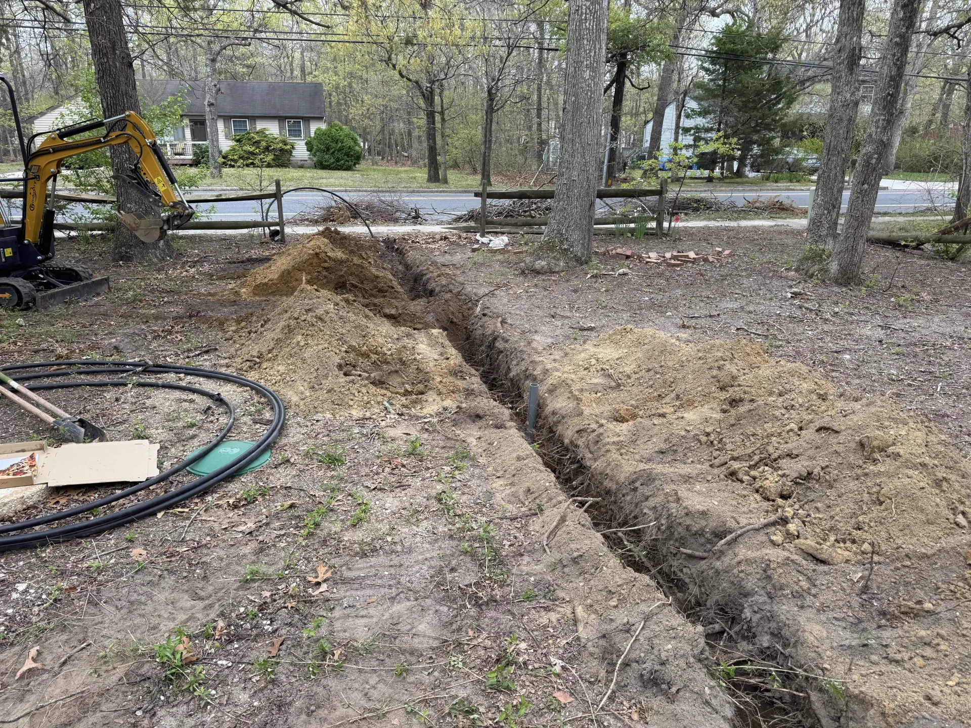 Excavator digging a trench beside two dirt piles in a wooded yard