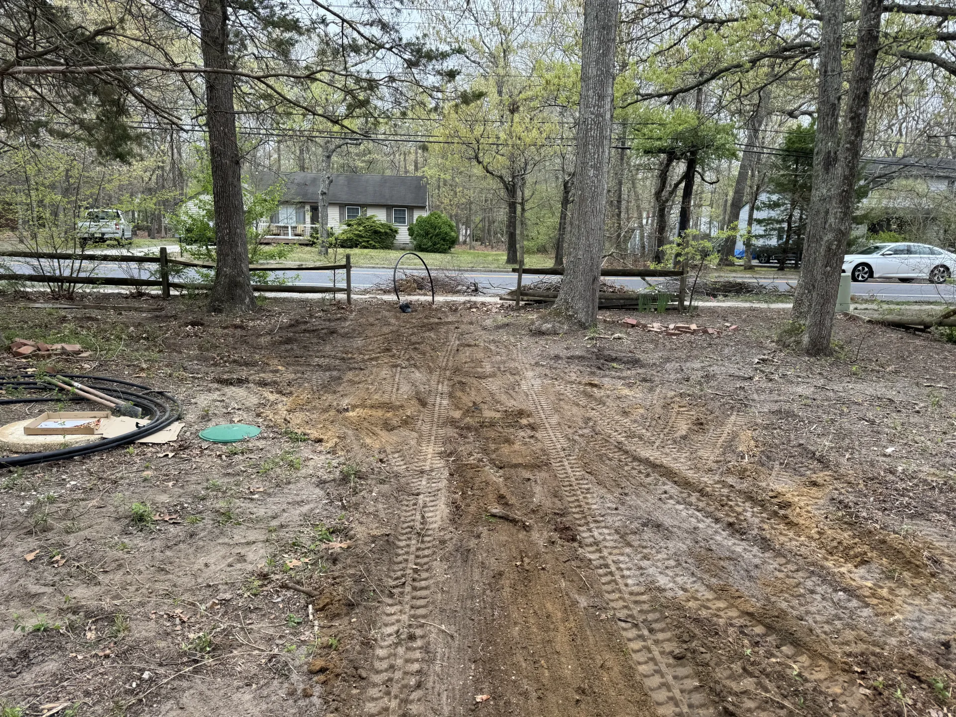 Dirt path through a wooded lot with trees and scattered leaves, near a road with parked cars.