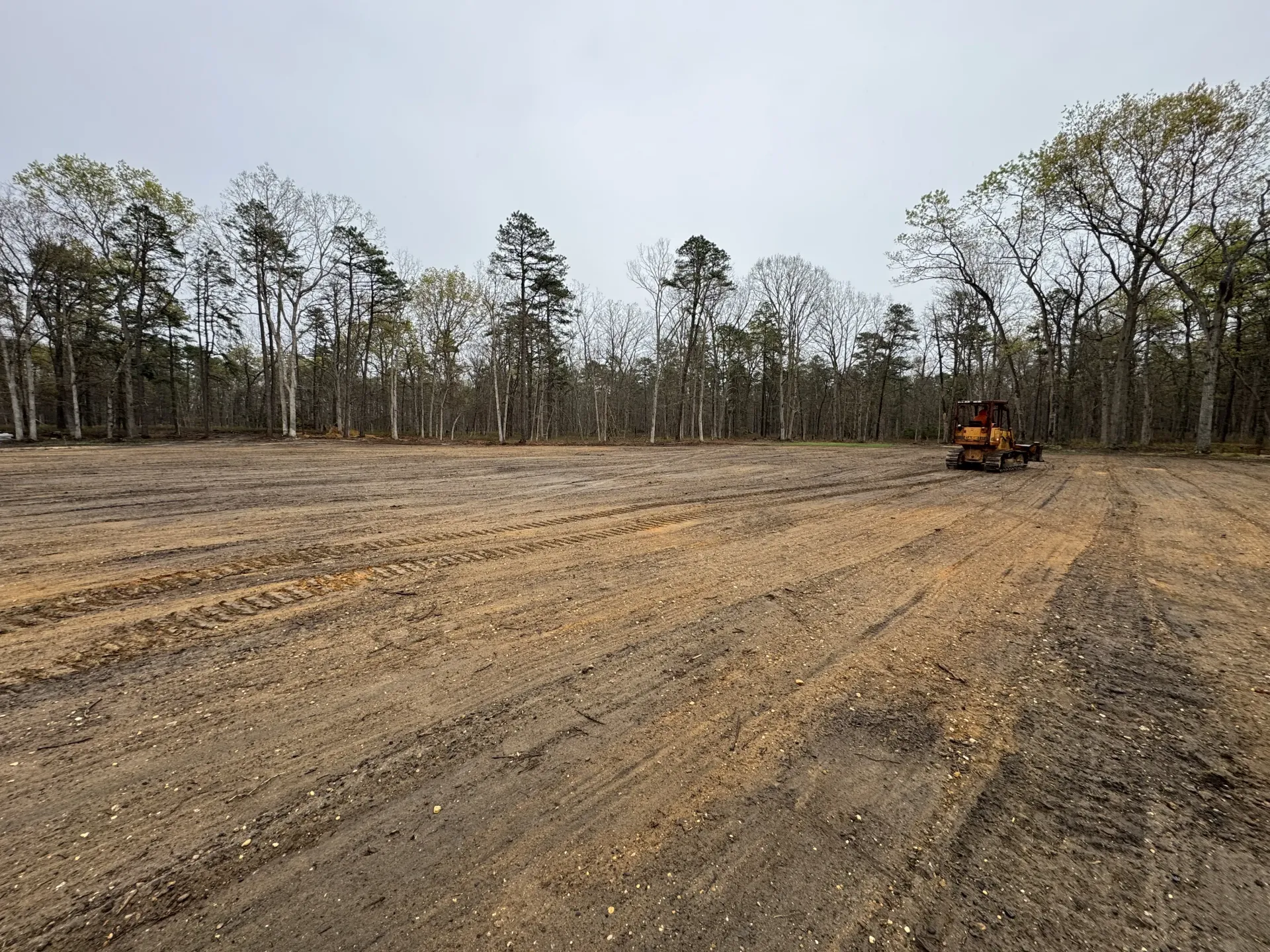 Cleared dirt field with tire tracks and an orange excavator near a tree line under a cloudy sky