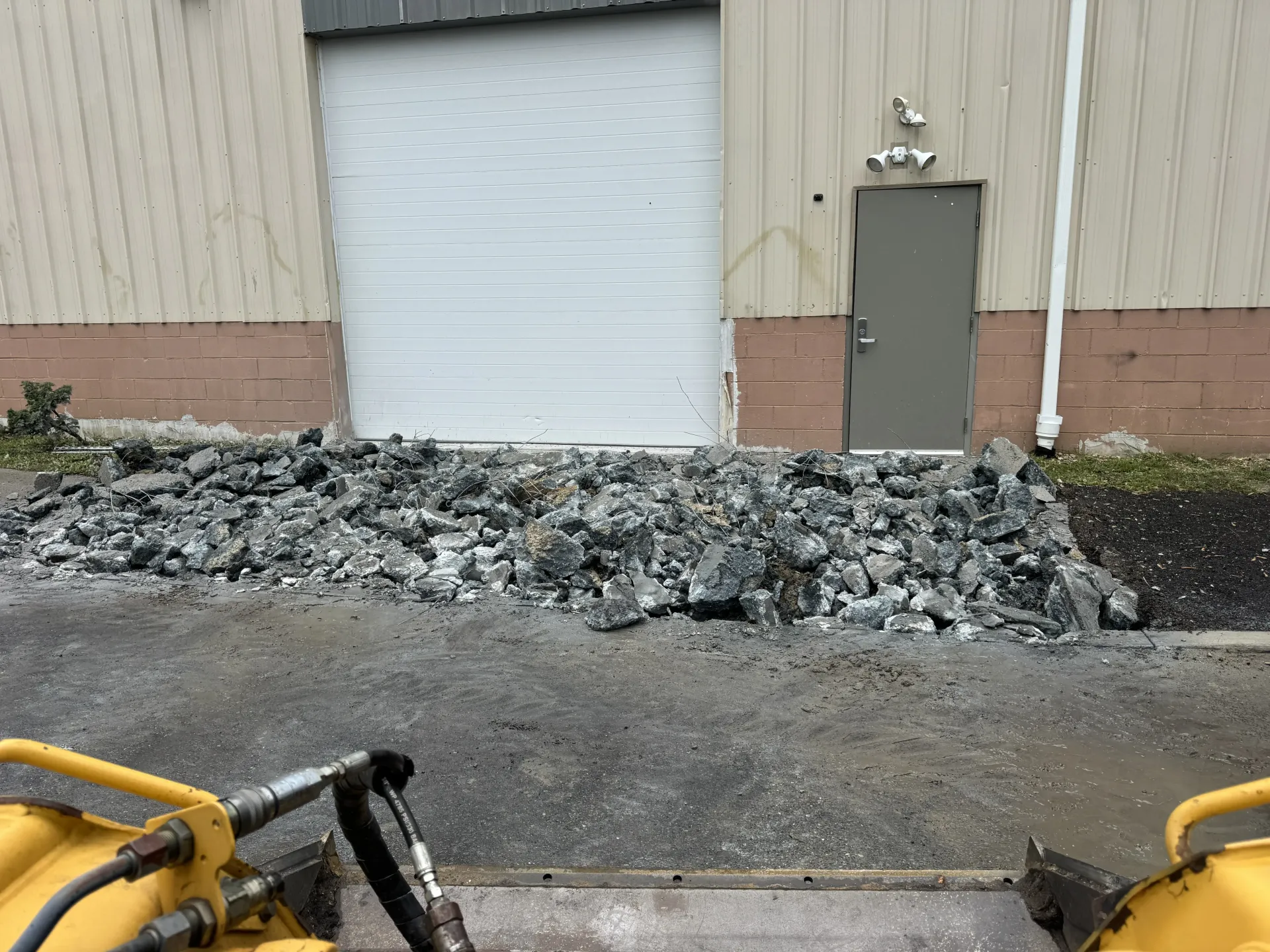 Industrial building exterior with a white roll-up door, gray side door, and pile of debris along the wall.