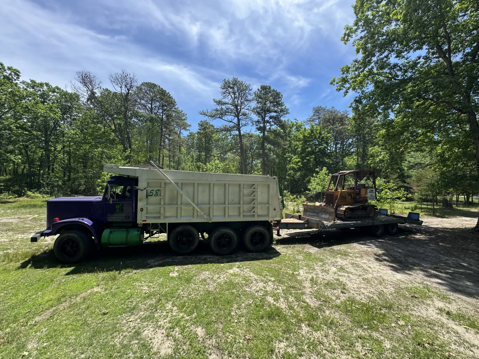 Purple truck hauling a backhoe on a flatbed trailer in a wooded clearing under a blue sky