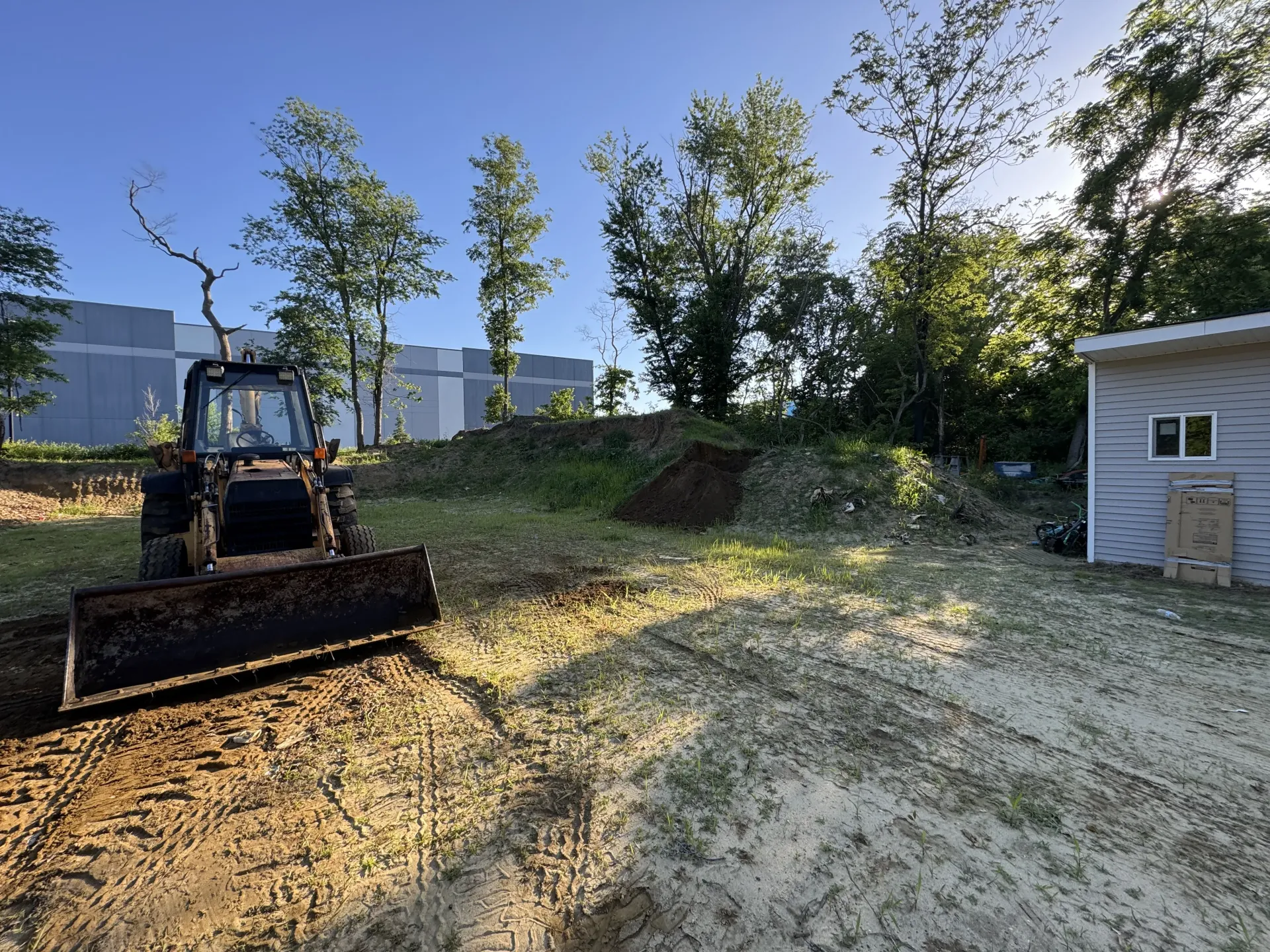 Bulldozer on dirt lot beside a small white shed, with trees and industrial buildings in the background.