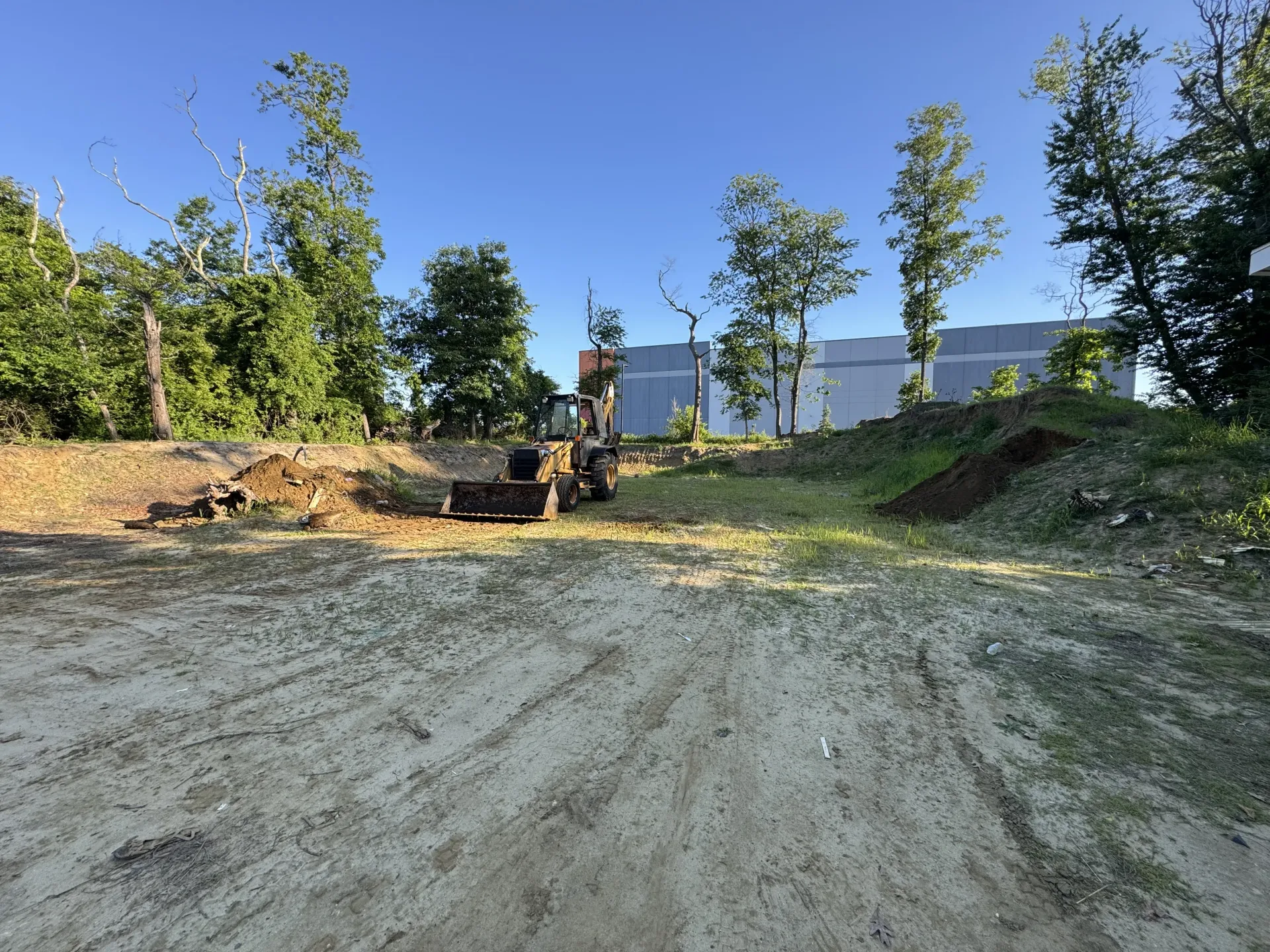 A person on a dirt bike rides up a gravel slope near trees and a building in bright sunlight.
